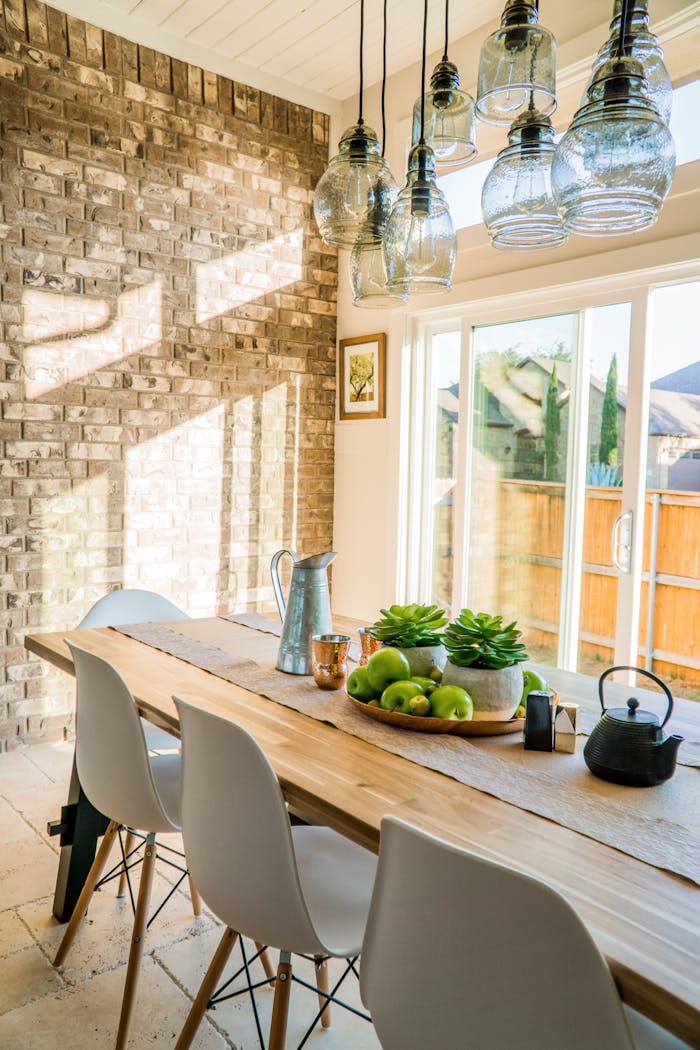 gallery-02 Sunlit dining room with contemporary decor and a rustic brick wall, featuring stylish lighting and a wooden table.
